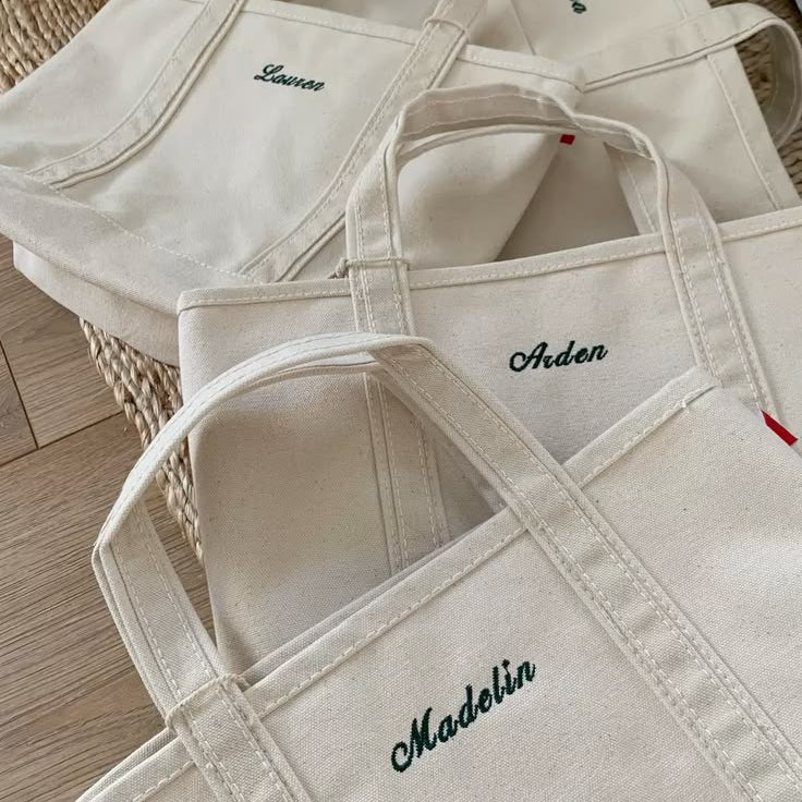 A group of off-white canvas tote bags with embroidered names including 'Madelin', 'Alden', and 'Lance', arranged on a light wood floor.