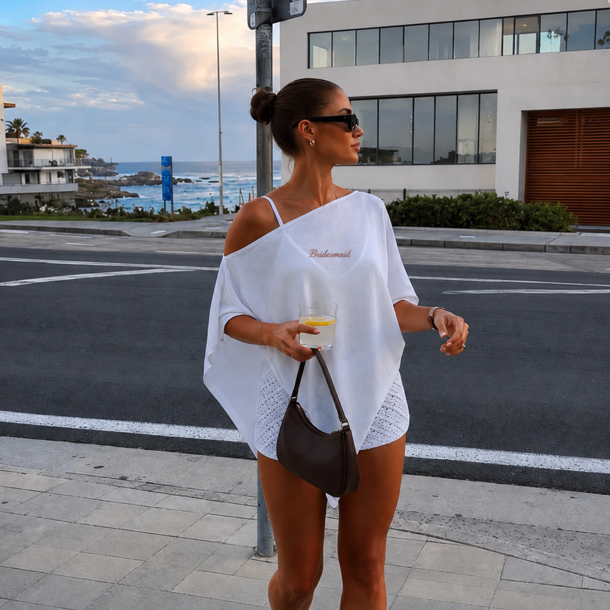 A woman stands on a city sidewalk by the sea, wearing a white one-shoulder crochet cover-up with 'Pandora' printed on the chest, holding a drink and carrying a dark brown shoulder bag.