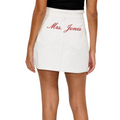 A model wearing a white mini skirt with 'Mrs. Jones' embroidered in red script on the back pocket, paired with a black top against a light gray background.