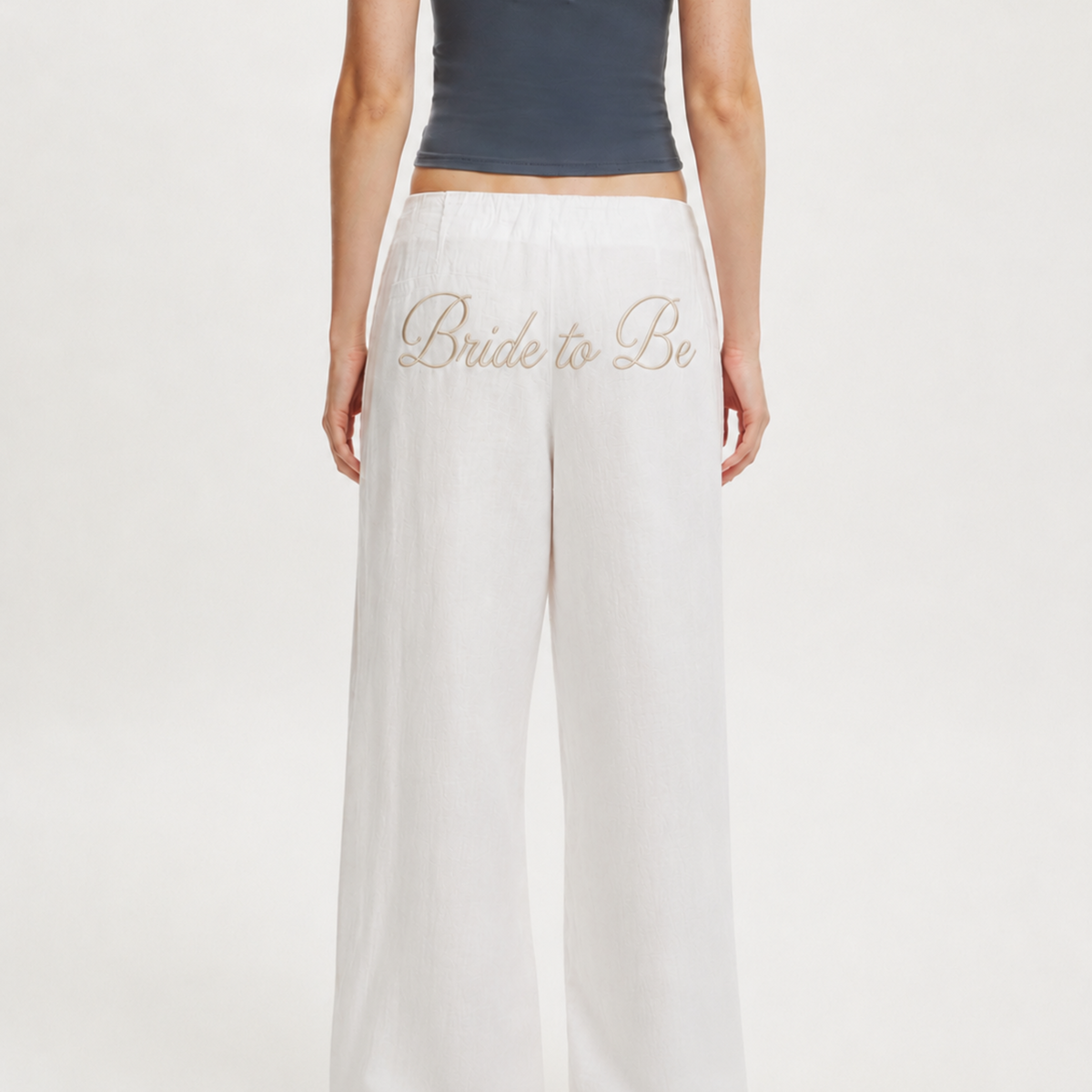 White wide-leg pants with 'Bride to Be' embroidered in gold cursive on the left thigh, shown on a model from the back against a light gray studio background.