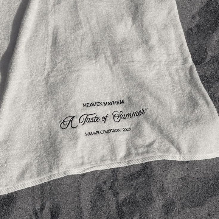 A close-up of a white fabric item, likely a towel or blanket, laid on dark textured ground. The fabric features black printed text reading 'HEAVEN MAYHEM', 'A Taste of Summer', and 'SUMMER COLLECTION 2023'.