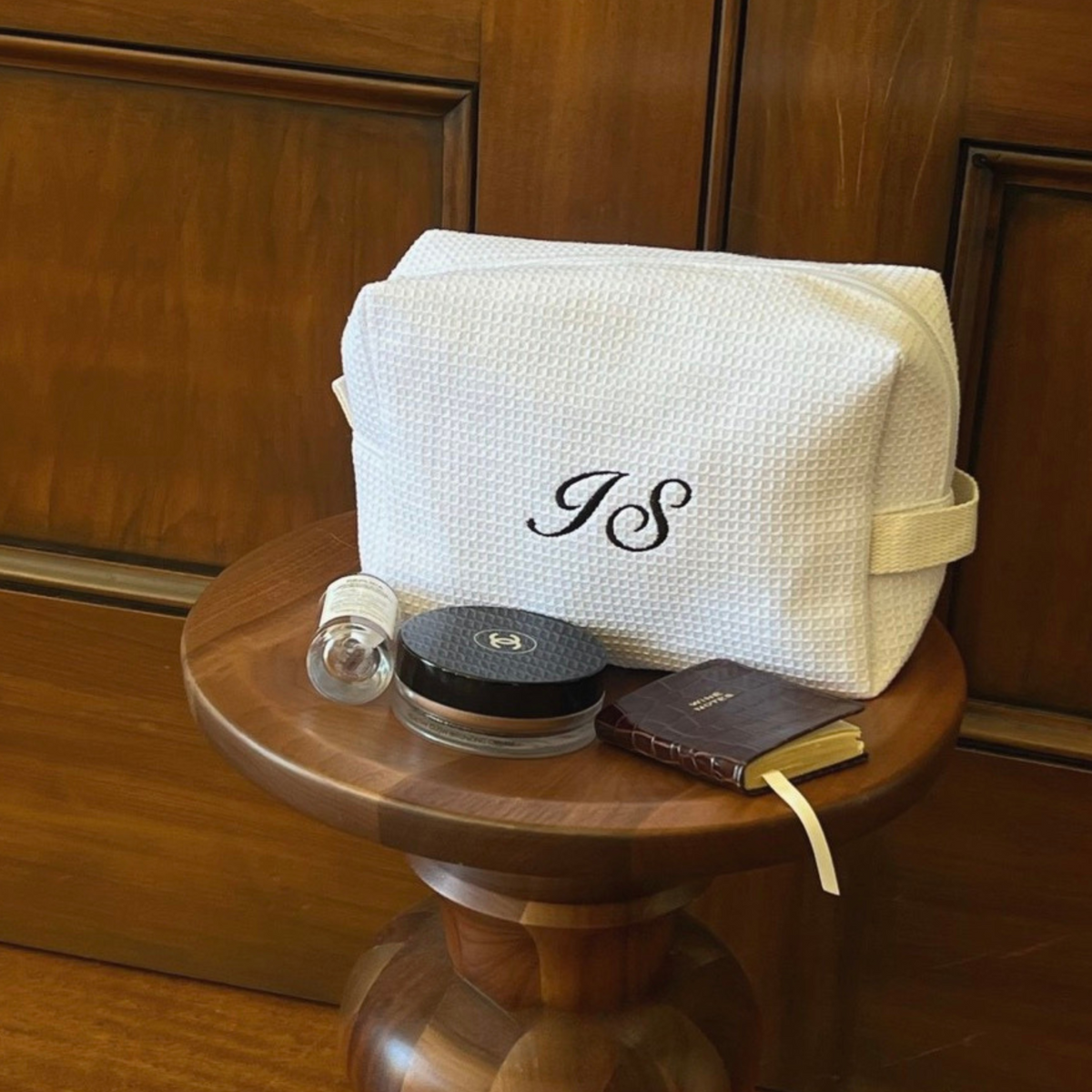 A cream-colored textured canvas cosmetic pouch with a black monogram 'JS' on the front, displayed on a round wooden side table alongside cosmetic items against a dark wood paneled background.