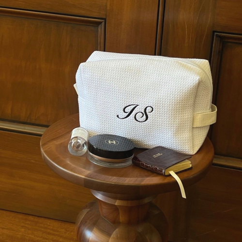 A cream-colored textured canvas cosmetic pouch with a black monogram 'JS' on the front, displayed on a round wooden side table alongside cosmetic items against a dark wood paneled background.