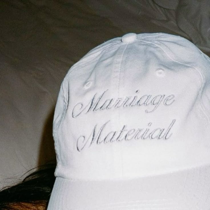 A white baseball cap with 'Marriage Material' embroidered in cursive script on the front, photographed close-up against a dark fabric background.