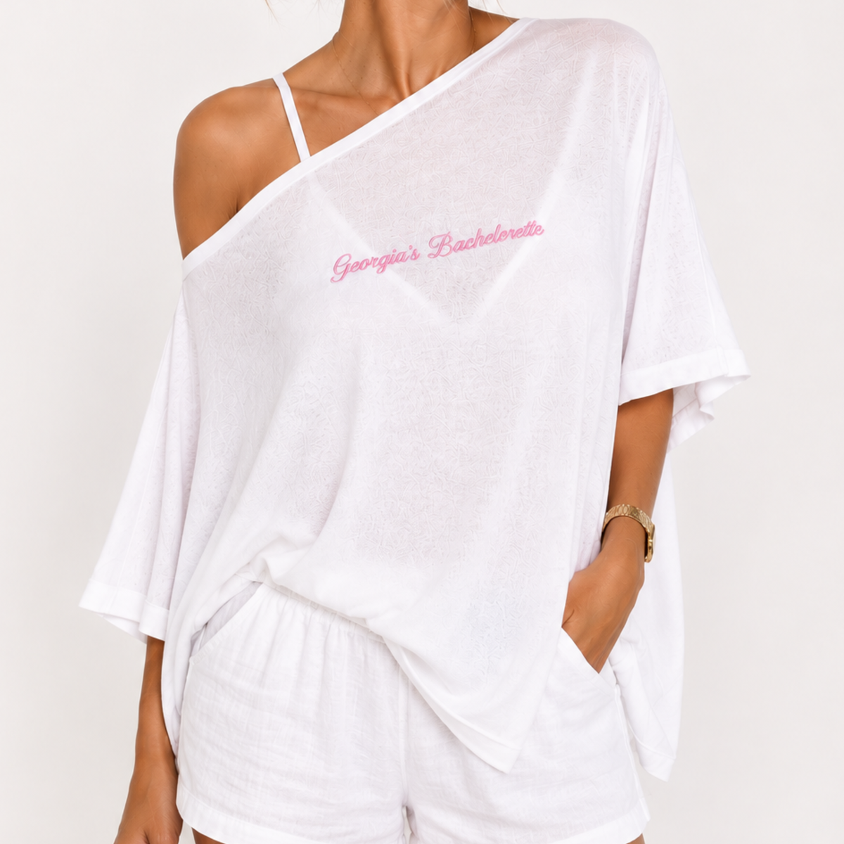 A woman wearing a white off-the-shoulder tunic top with pink cursive embroidery reading 'Georgia's Bachelorette' on the chest, paired with white shorts, against a plain light background.