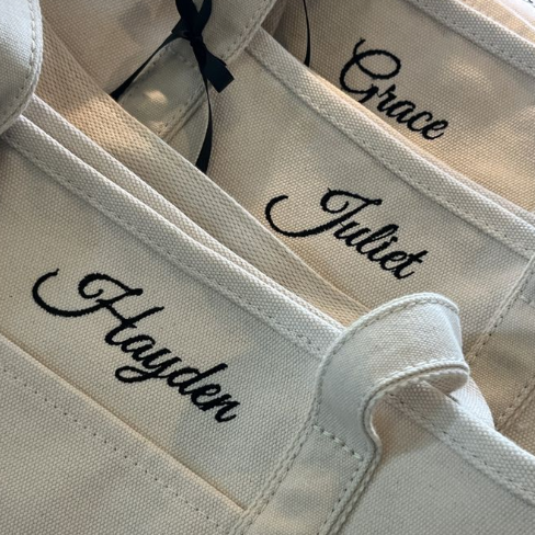 Close-up of multiple cream canvas tote bags with black embroidered names, including Grace, Juliet, and Hayden, shown on a neutral background.