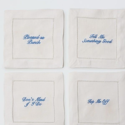 Four square off-white napkins arranged in a 2x2 grid on a white background, each with a stitched border and blue embroidered phrase: 'Pleased as Punch,' 'Tell Me Something Good,' 'Don't Mind If I Do,' and 'Top Me Off.'.