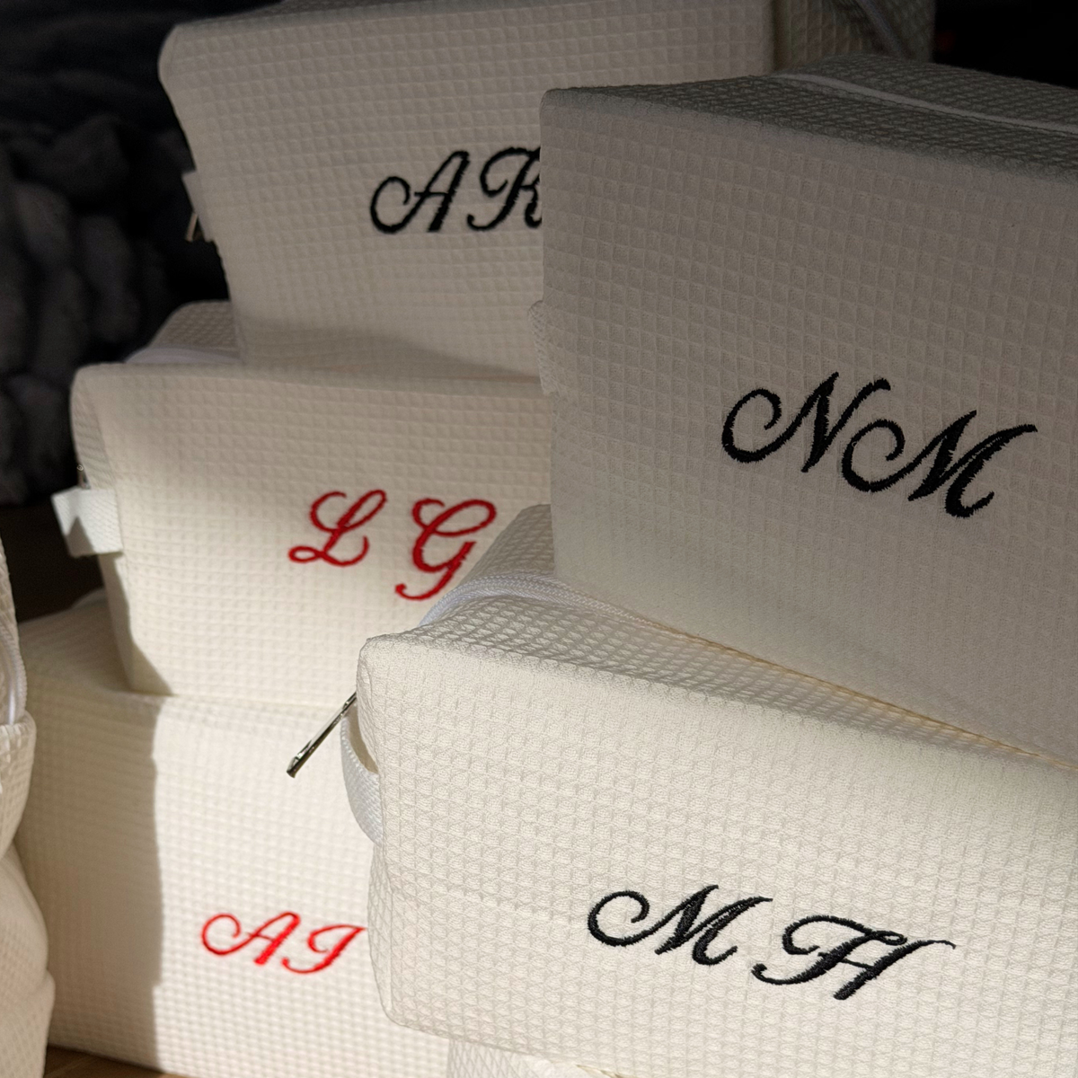 A stack of white waffle-weave zippered pouches or small cases, each personalized with embroidered initials in black or red script. The initials visible include NM, MH, LG, and AI.