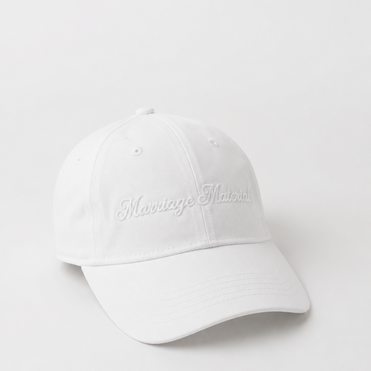A white baseball cap with 'Marriage Material' embroidered in white script on the front, photographed on a seamless white background.