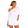 A woman modeling a white two-piece lounge set consisting of an asymmetrical off-the-shoulder top with pink script text and matching white shorts, set against a plain white background.